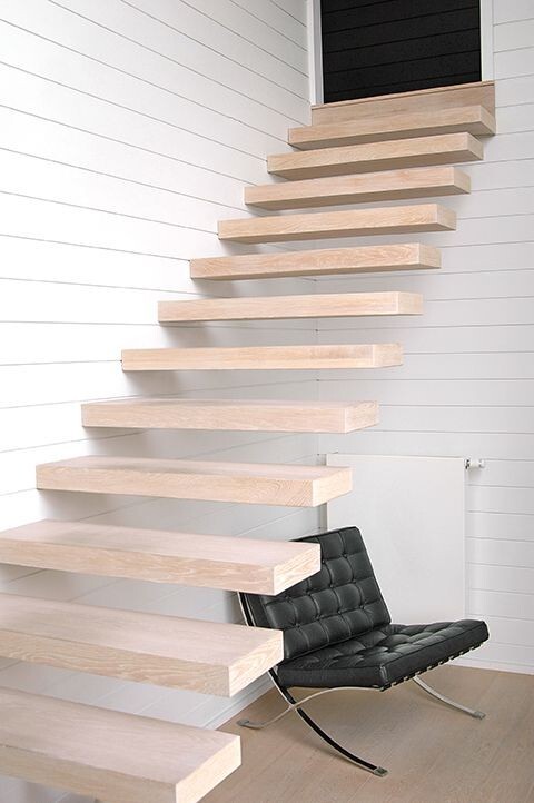 Moderne zwevende trap met lichte houten treden tegen een witte lamellenwand, met een zwarte designstoel op een lichtgrijze vloer.