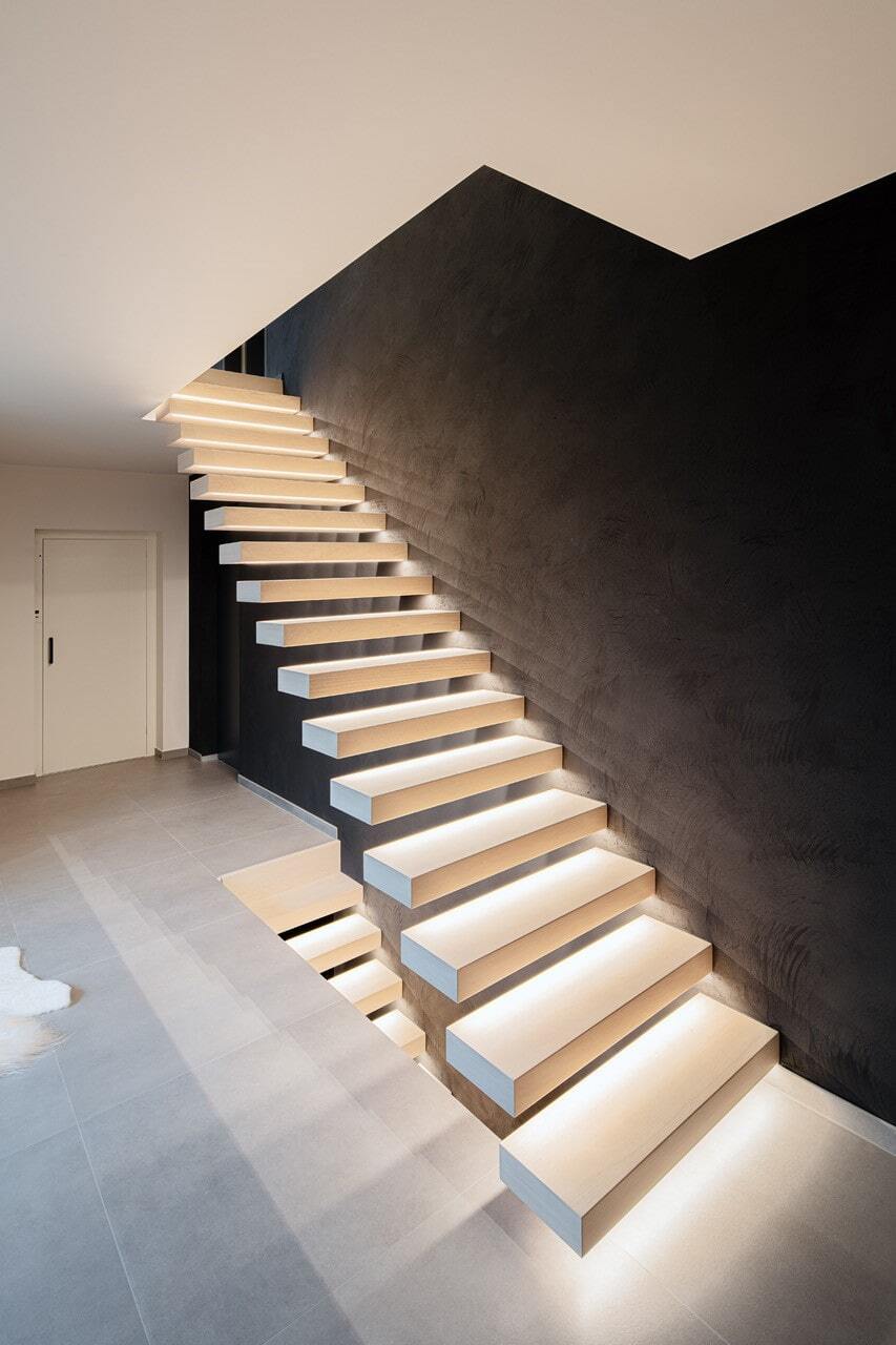Moderne zwevende trap met lichte treden en subtiele ledverlichting onder elke trede, geplaatst tegen een donkere muur in een strak vormgegeven interieur.