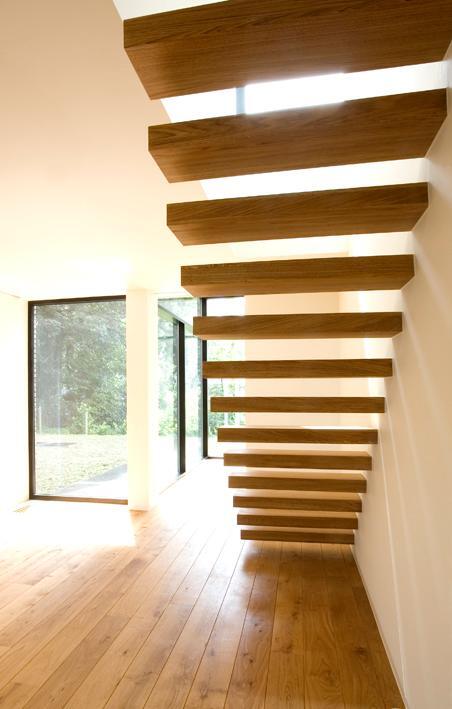 Onderzijde van een moderne zwevende trap met dikke houten treden aan een witte muur, boven een houten vloer in een lichte ruimte met grote ramen.