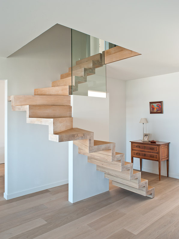 Architectonische trap in licht hout met zigzagvorm en glazen balustrade, geplaatst in een helder interieur met houten vloer, witte muren en een klassiek houten ladekastje.